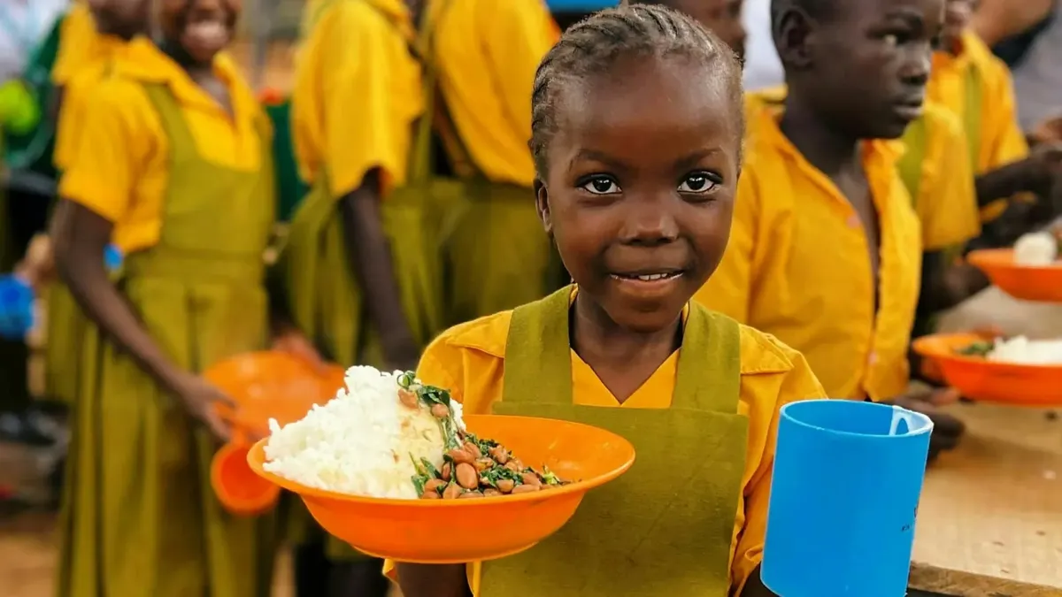 A schoolgirl in a yellow uniform smiles as she holds an orange bowl of rice and beans